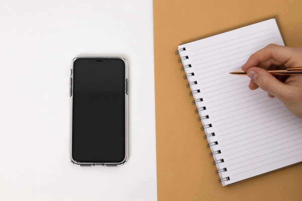 A smartphone beside a notebook as a person writes with a pen