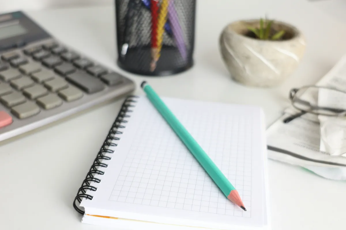 Calculator, squared notebook and teal pencil on a desk beside glasses and a small plant.