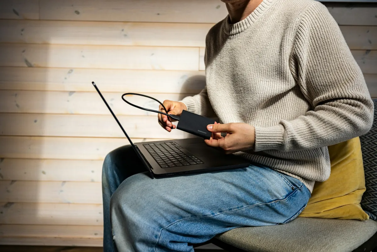 Person holding a black portable storage drive and cable above a laptop on their lap.