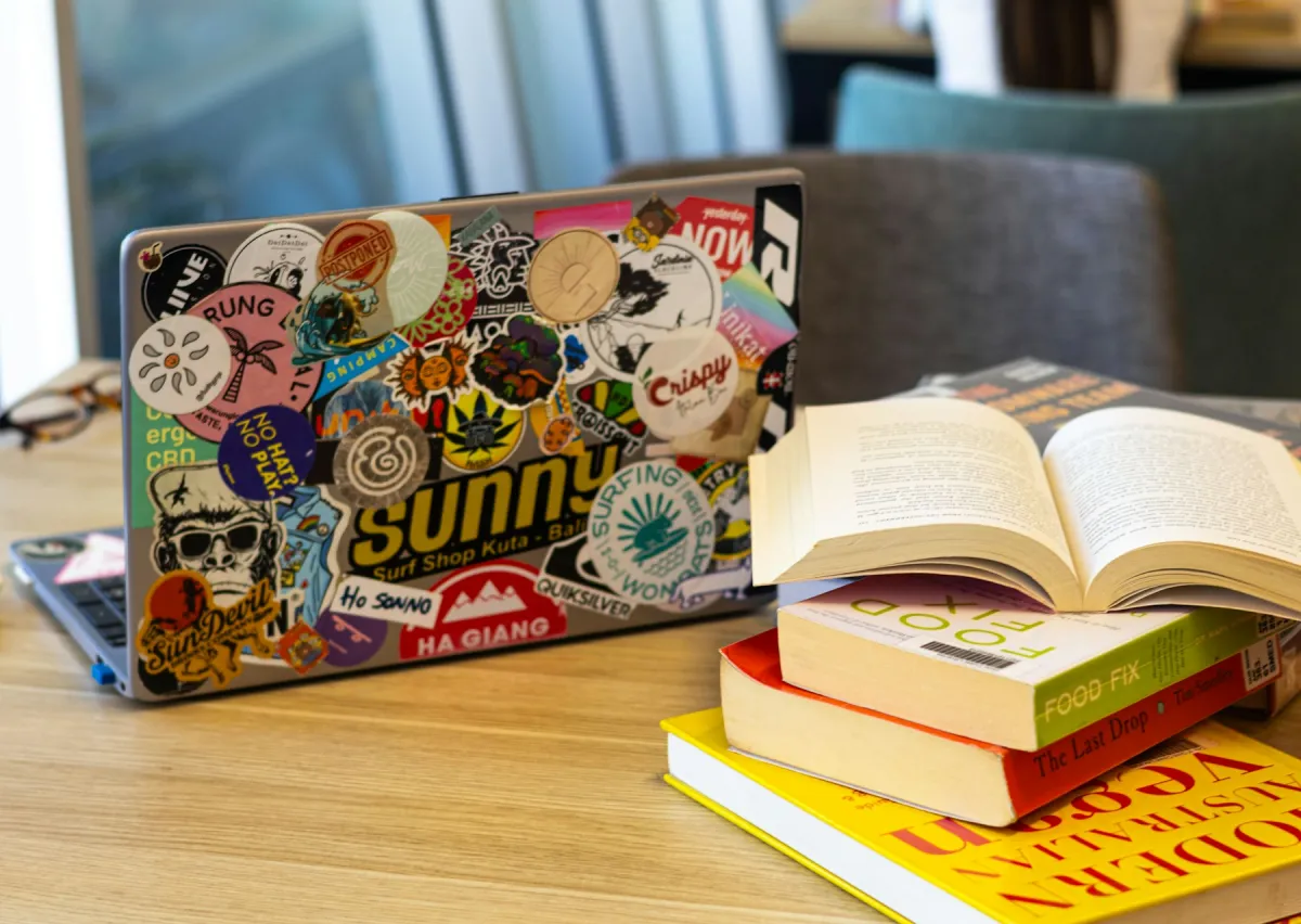 Sticker-covered laptop beside an open book and a stack of books on a wooden table.