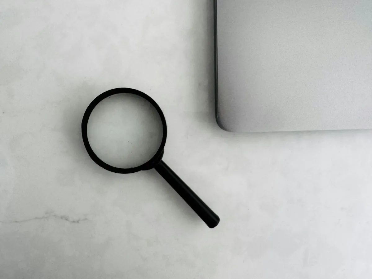 Black magnifying glass beside the corner of a silver laptop on a pale marble surface.