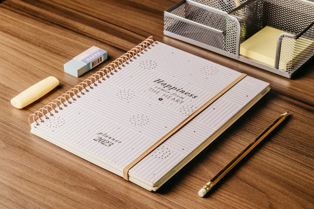 Pink spiral planner, pencil, highlighter, eraser and metal stationery organizer on a wooden desk.