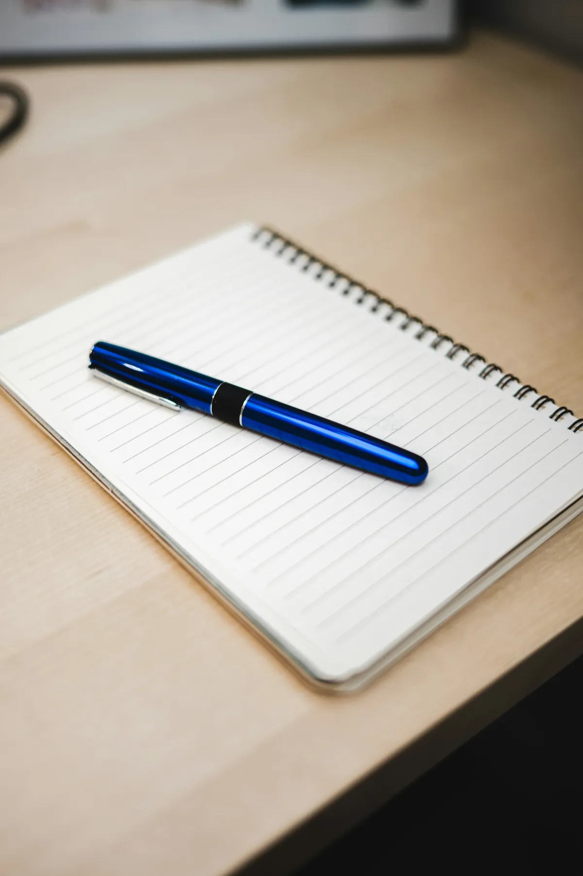 Blue fountain pen resting diagonally on a blank lined spiral notebook on a wooden desk.