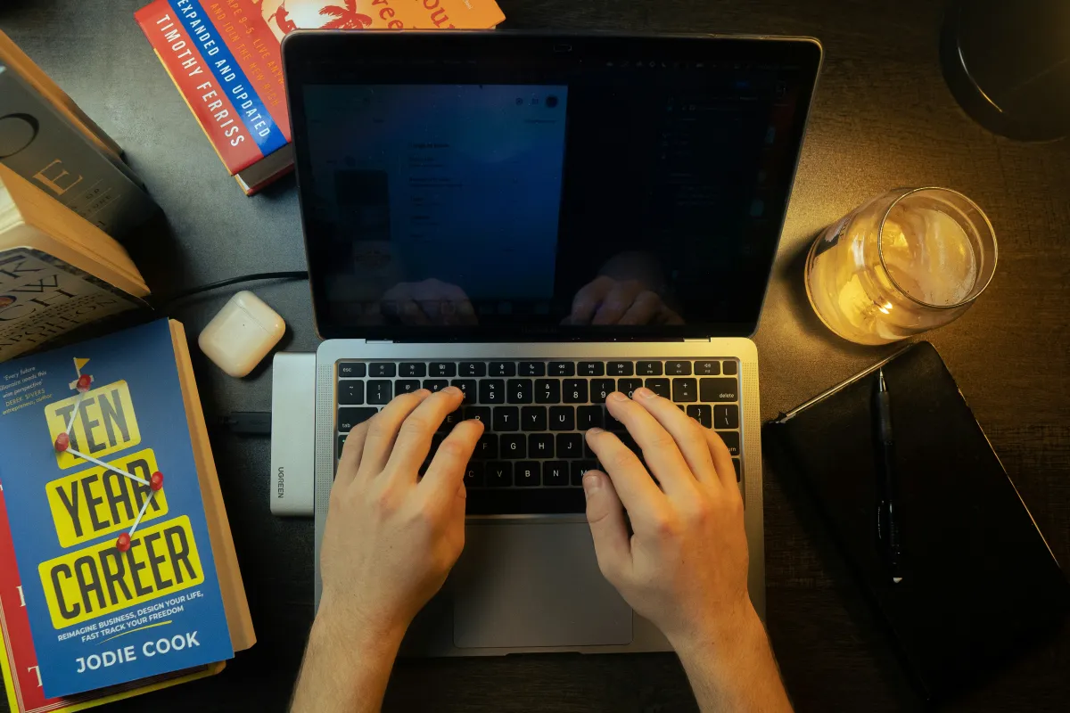 Hands typing on a laptop surrounded by books, a notebook and a drink on a wooden desk.
