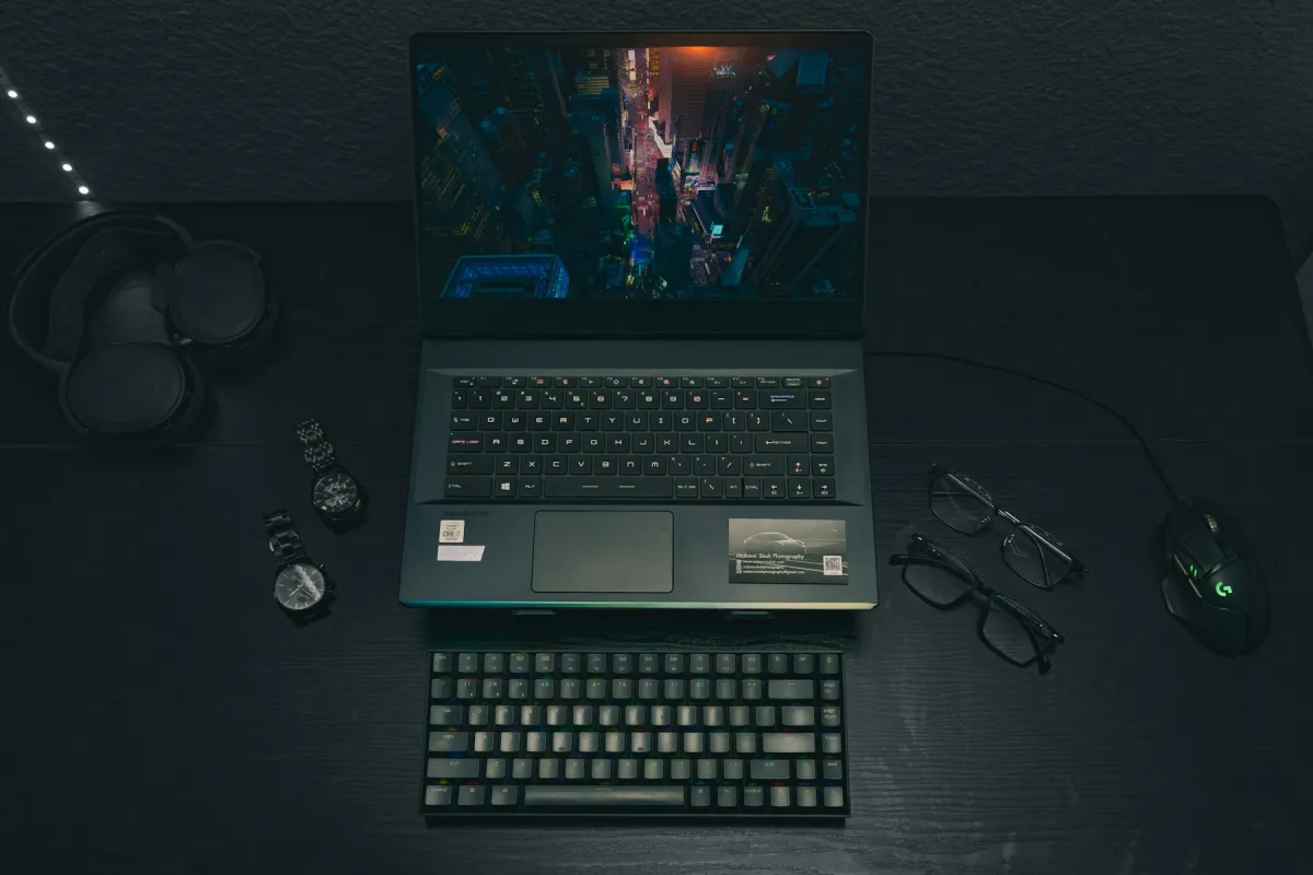 Top-down photograph of a gaming laptop, keyboard, headphones, watches, glasses and mouse on a dark desk.