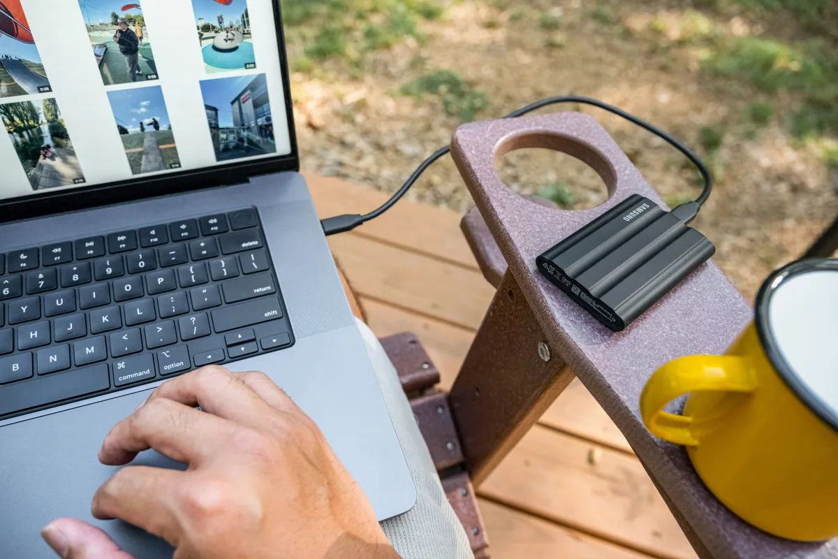 A person uses a laptop outdoors with a black Samsung portable drive connected beside a yellow mug.