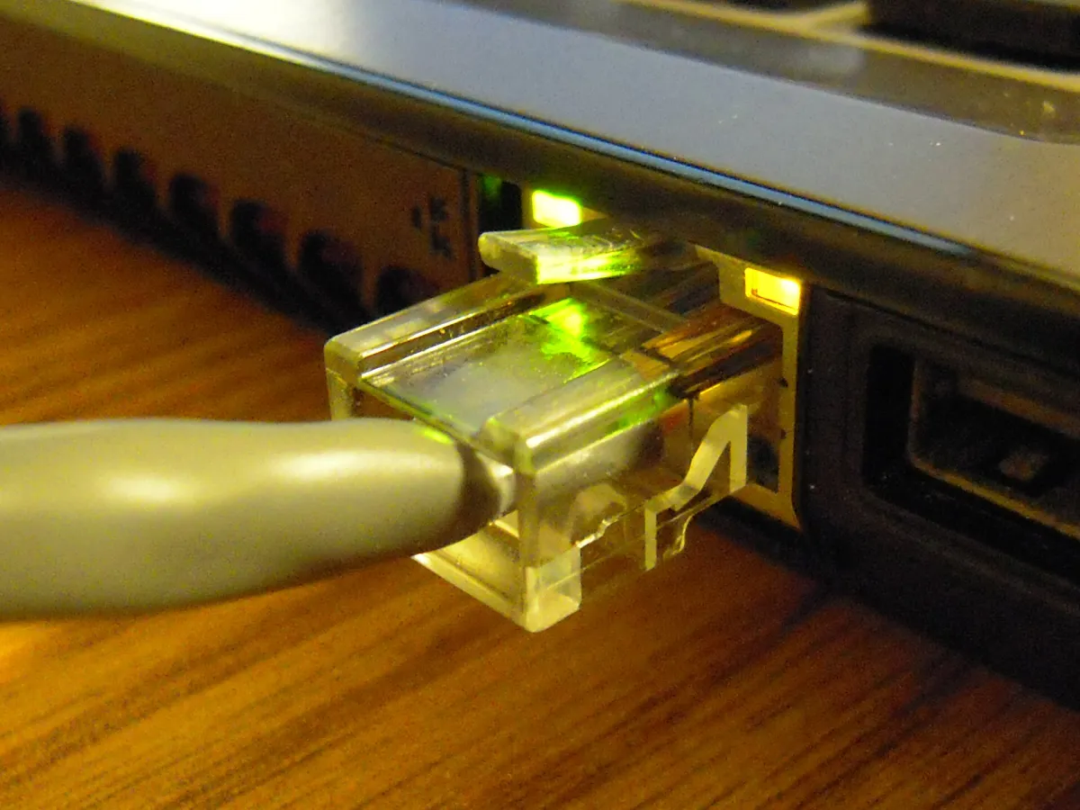 Close-up of a clear Ethernet plug connected to a laptop, with green and yellow lights beside the socket.