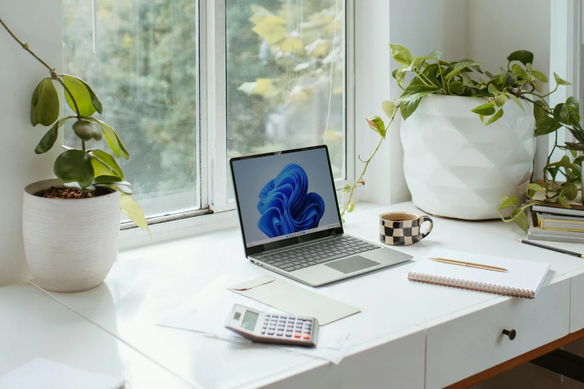Open Windows laptop on a white desk by a window, with plants, a mug, notebook and calculator.