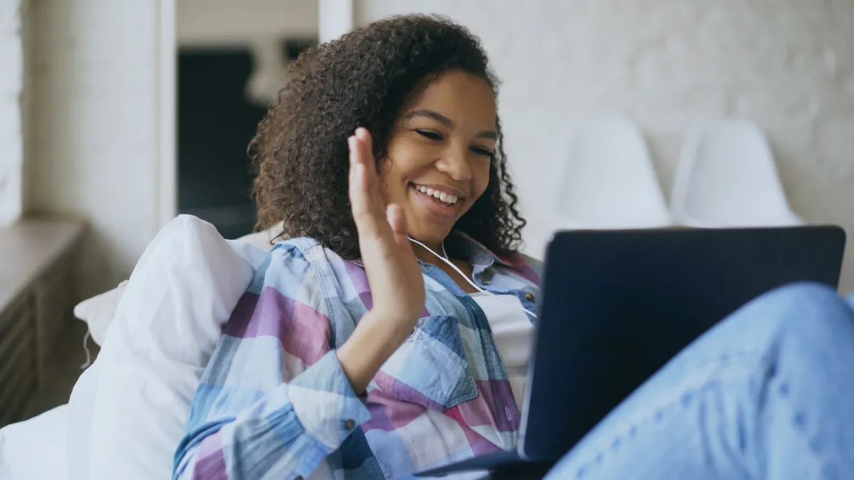 Woman wearing wired earphones smiling and waving toward a laptop at home.