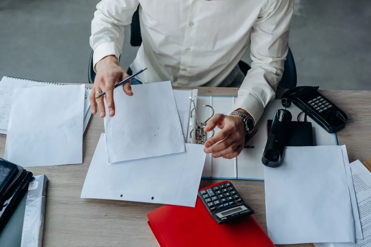 Person sorting loose papers into a ring binder at a desk with a calculator and telephone.