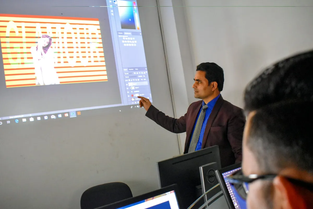 Instructor pointing at a projected software demonstration in a classroom with computer monitors in the foreground.