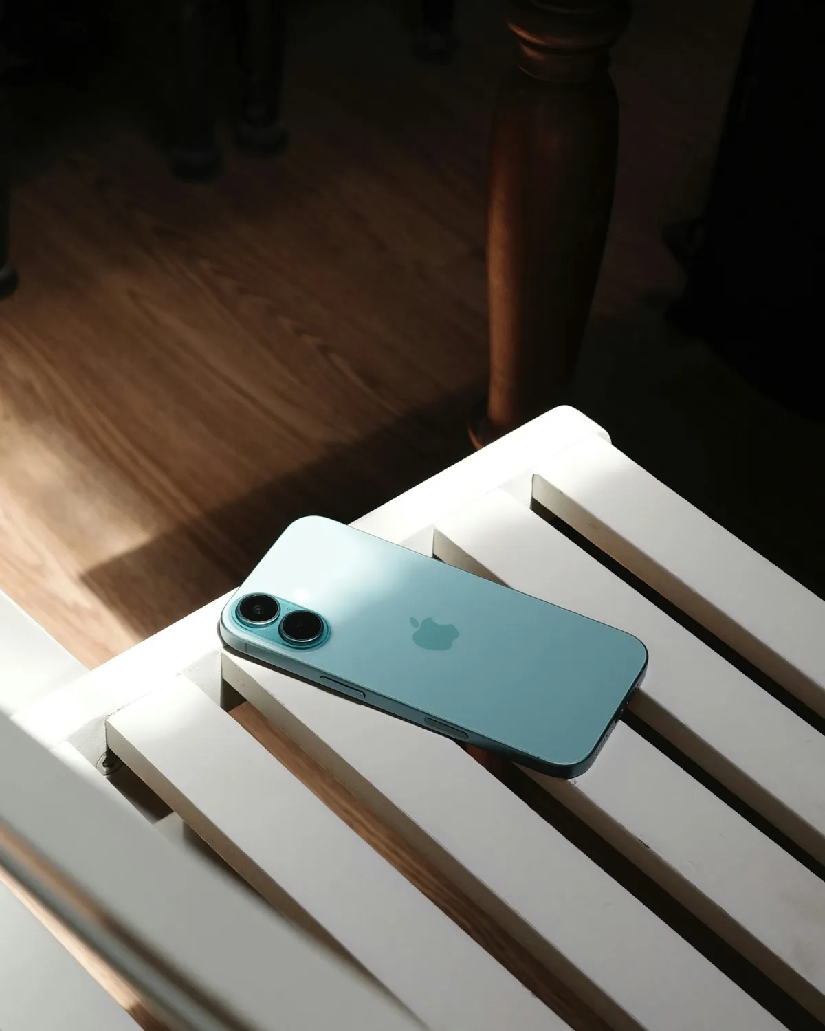 A teal iPhone lying face down on a white slatted wooden bench in sunlight.