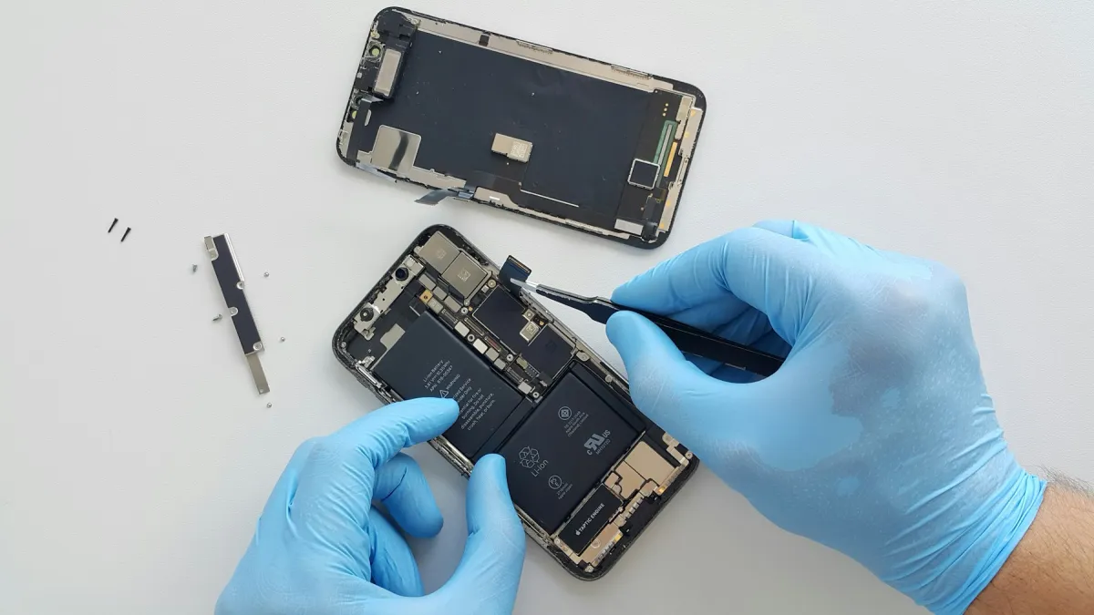 Blue-gloved hands use tweezers on an opened iPhone beside its detached display on a white repair bench.
