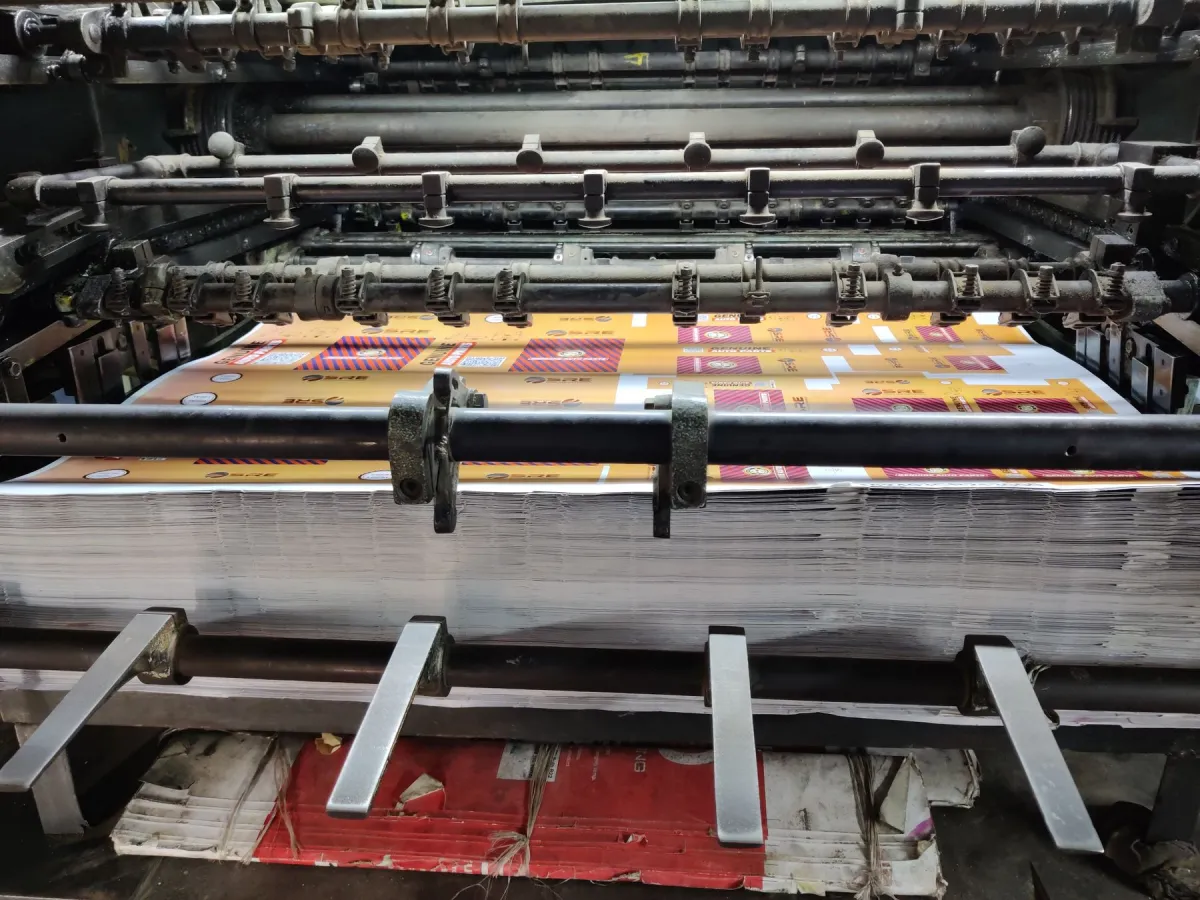 Stack of colorful printed sheets beneath the metal rollers and guides of an industrial printing machine.