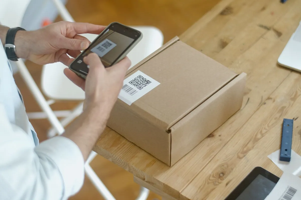 Hands holding a smartphone over a QR-labelled cardboard parcel on a wooden table.