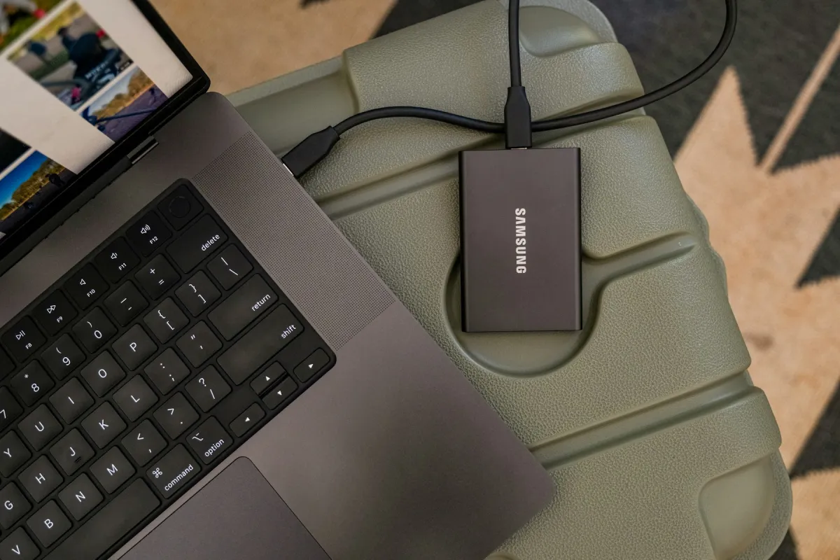 Portable Samsung storage drive connected by cable to a laptop on a green chair.