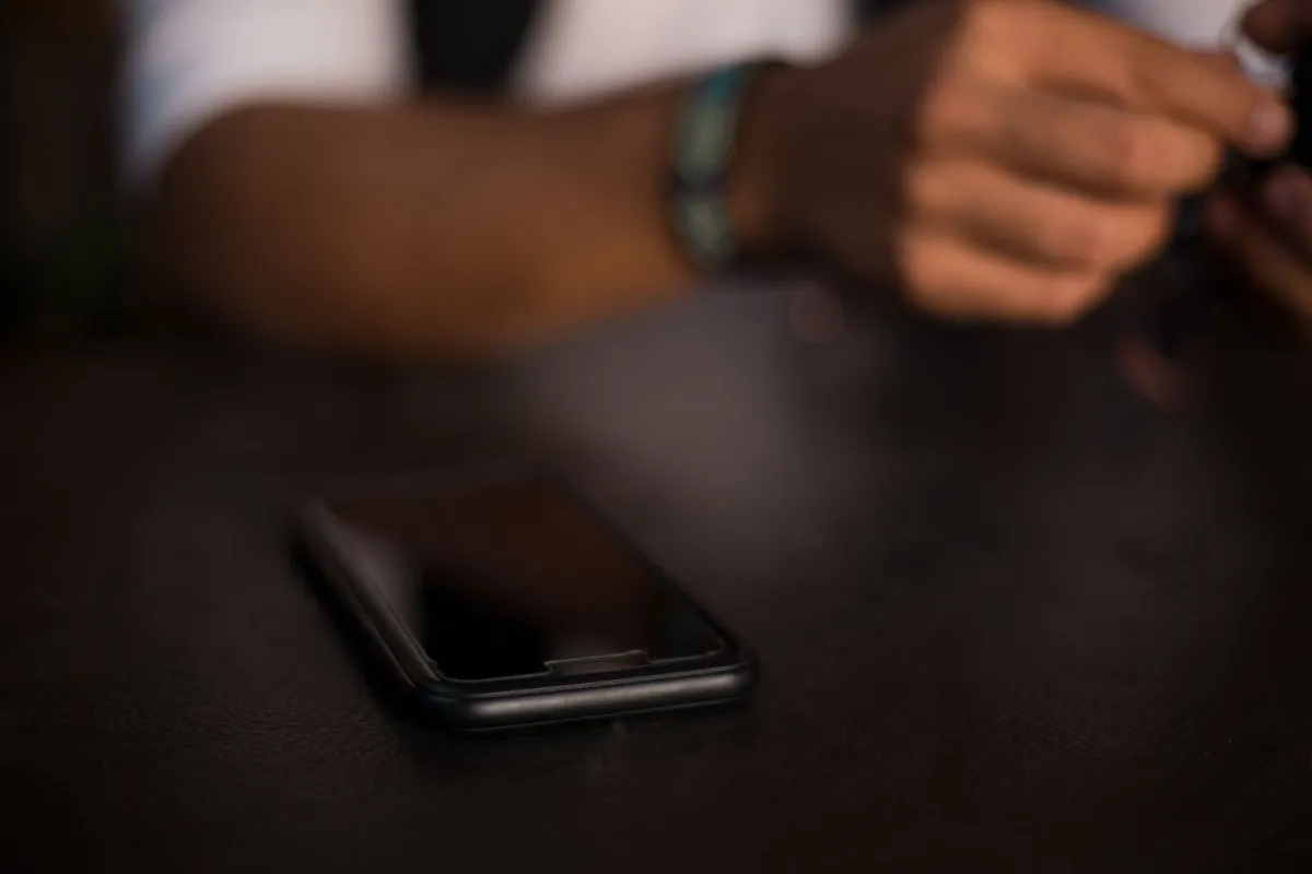A smartphone with a dark reflective screen lies face-up on a table, with a person’s hands blurred in the background.