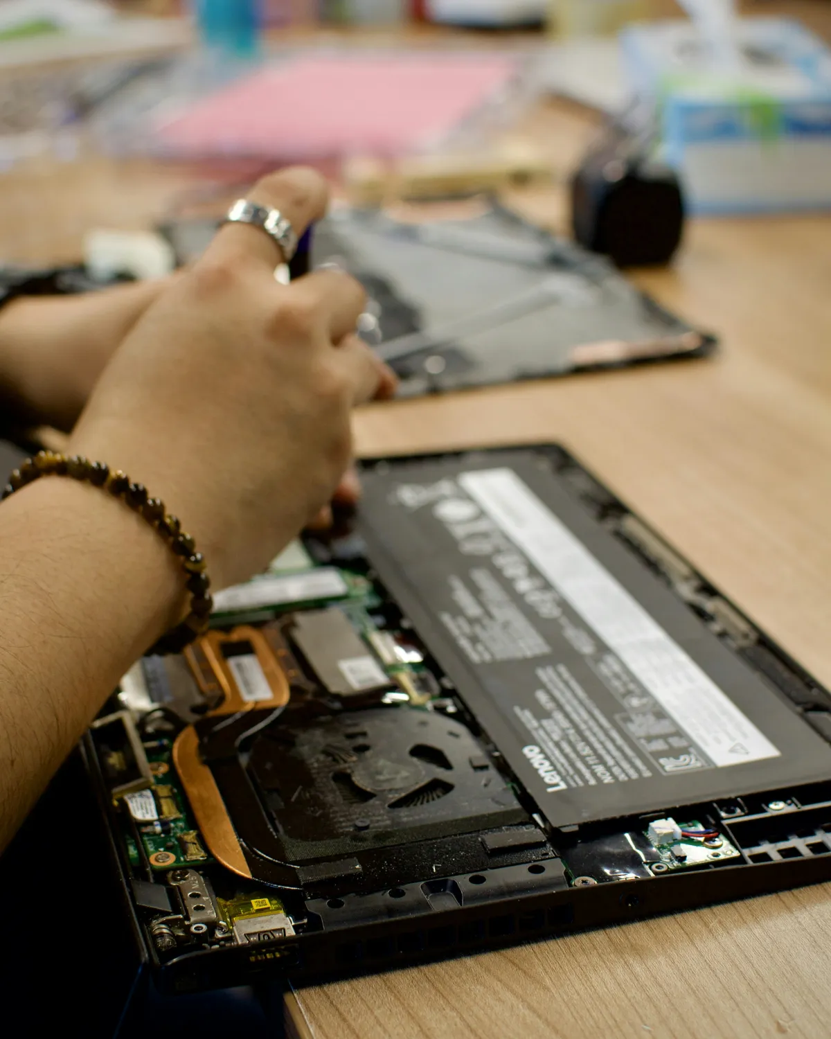 Hands working over an opened Lenovo laptop on a wooden desk, with its internal battery, fan and copper heat pipes visible.