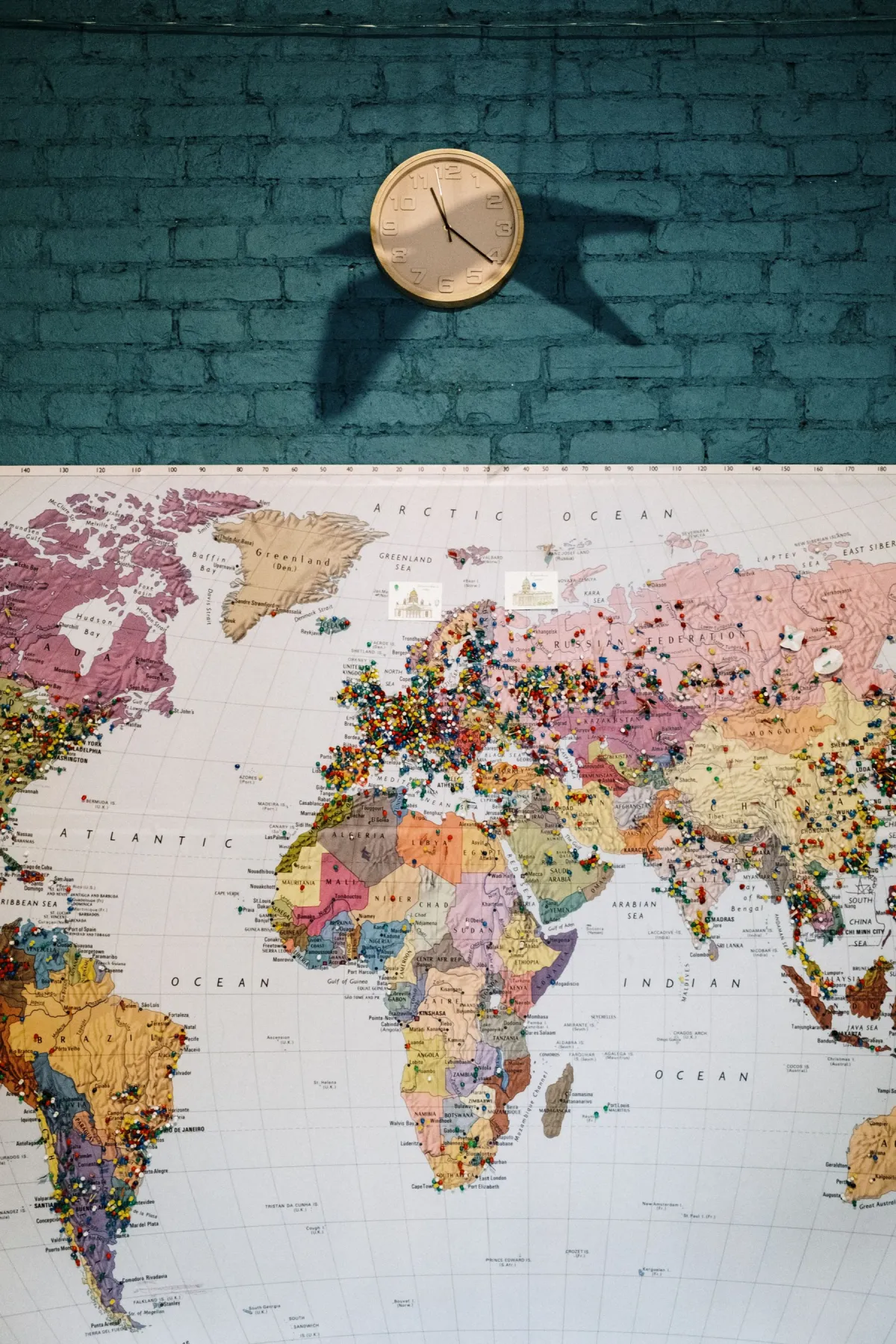 Analog wall clock above a world map covered with colored pins on a blue brick wall.