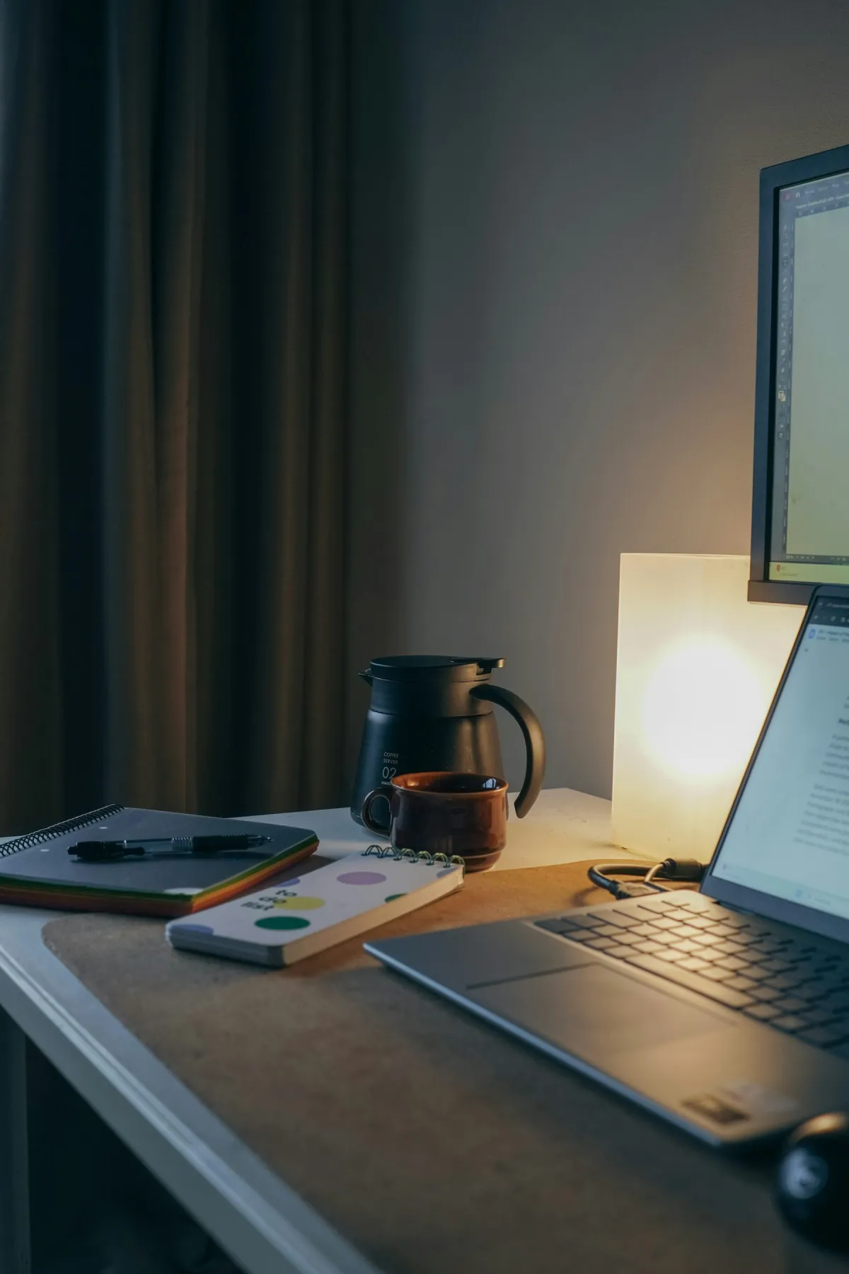 Laptop and monitor on a home desk beside two notebooks, a pen, coffee cup and coffee server.