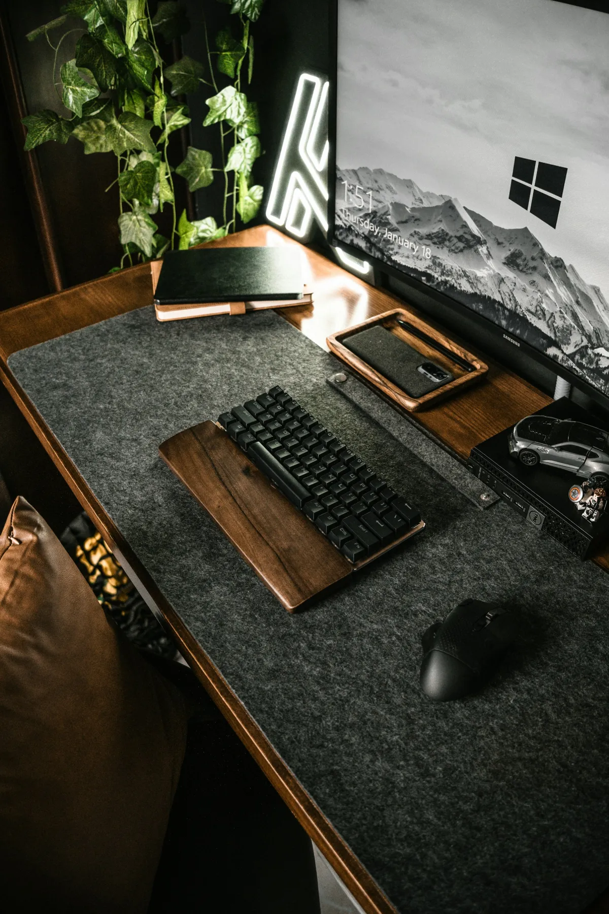 Compact black keyboard with a wooden palm rest and black mouse on a desk mat beside a monitor and notebooks.
