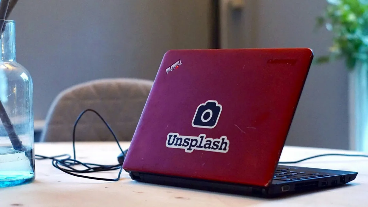 A red Lenovo ThinkPad with a camera and Unsplash sticker on its lid, open on a wooden table beside a glass bottle.