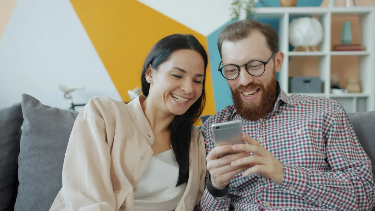 Two adults sitting together on a couch at home, smiling as they look at a smartphone.