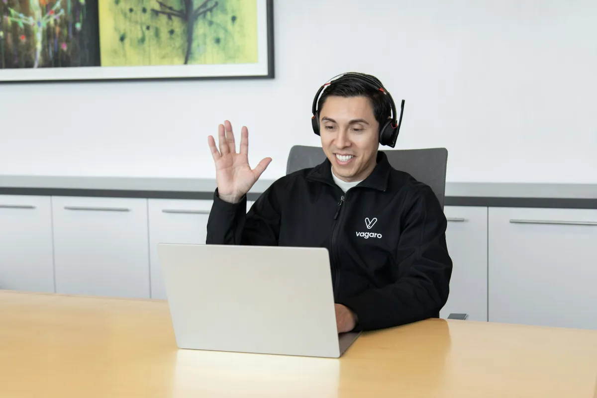 A man wearing a headset waves while seated at a laptop in an office.