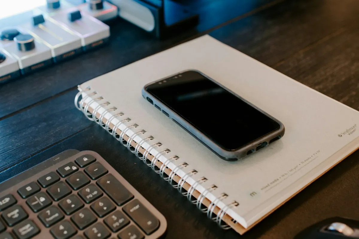 A phone with a dark screen rests on a spiral notebook beside computer keyboards.