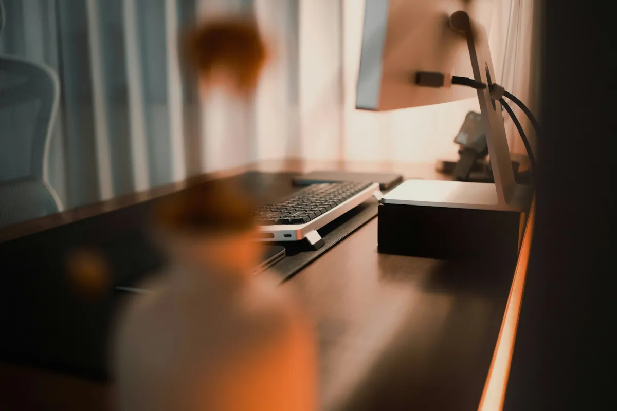 Side and rear view of a desktop monitor on a stand, with connected cables and a keyboard on a warmly lit desk.