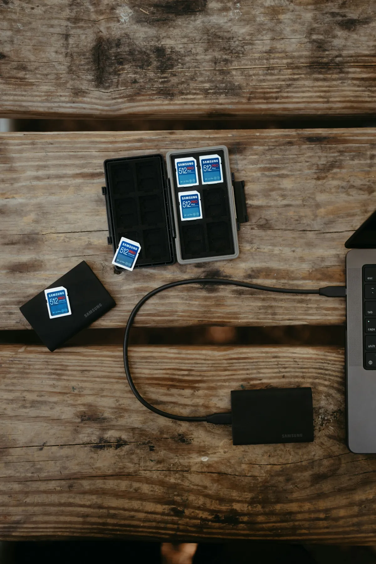 SD memory cards in an open case and two black external storage devices beside the edge of a laptop on a wooden table.
