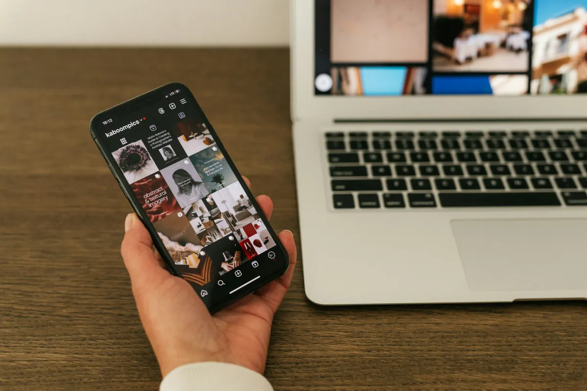 A hand holds a phone displaying a social profile beside an open laptop on a wooden desk.
