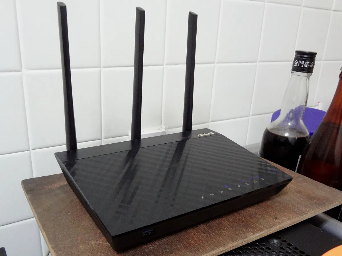 Black ASUS RT-AC66U B1 router with three upright antennas on a wooden shelf.