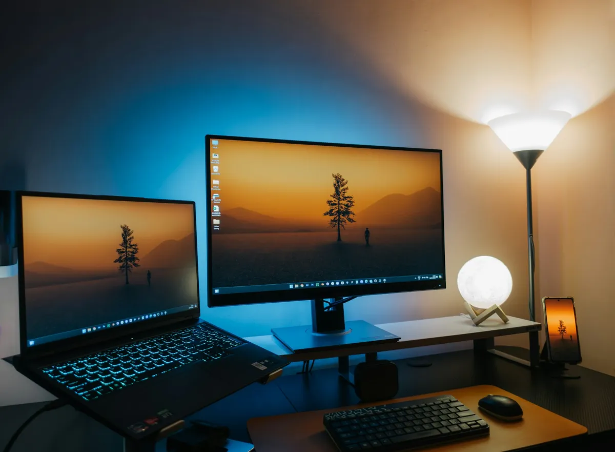 Laptop beside a larger external monitor, with matching landscape wallpapers, a keyboard and desk lamps.