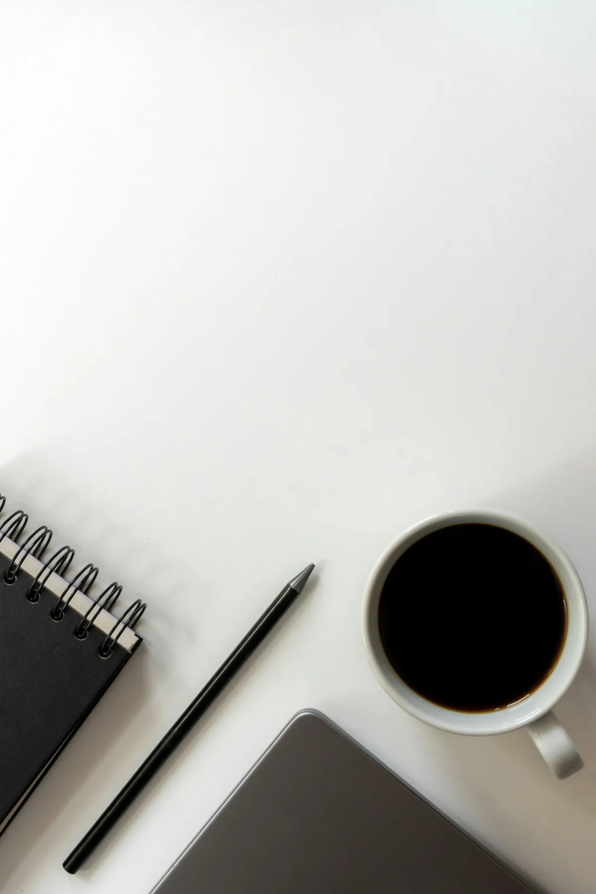 Coffee, a black spiral notebook and pencil beside the corner of a closed laptop on a white desk.