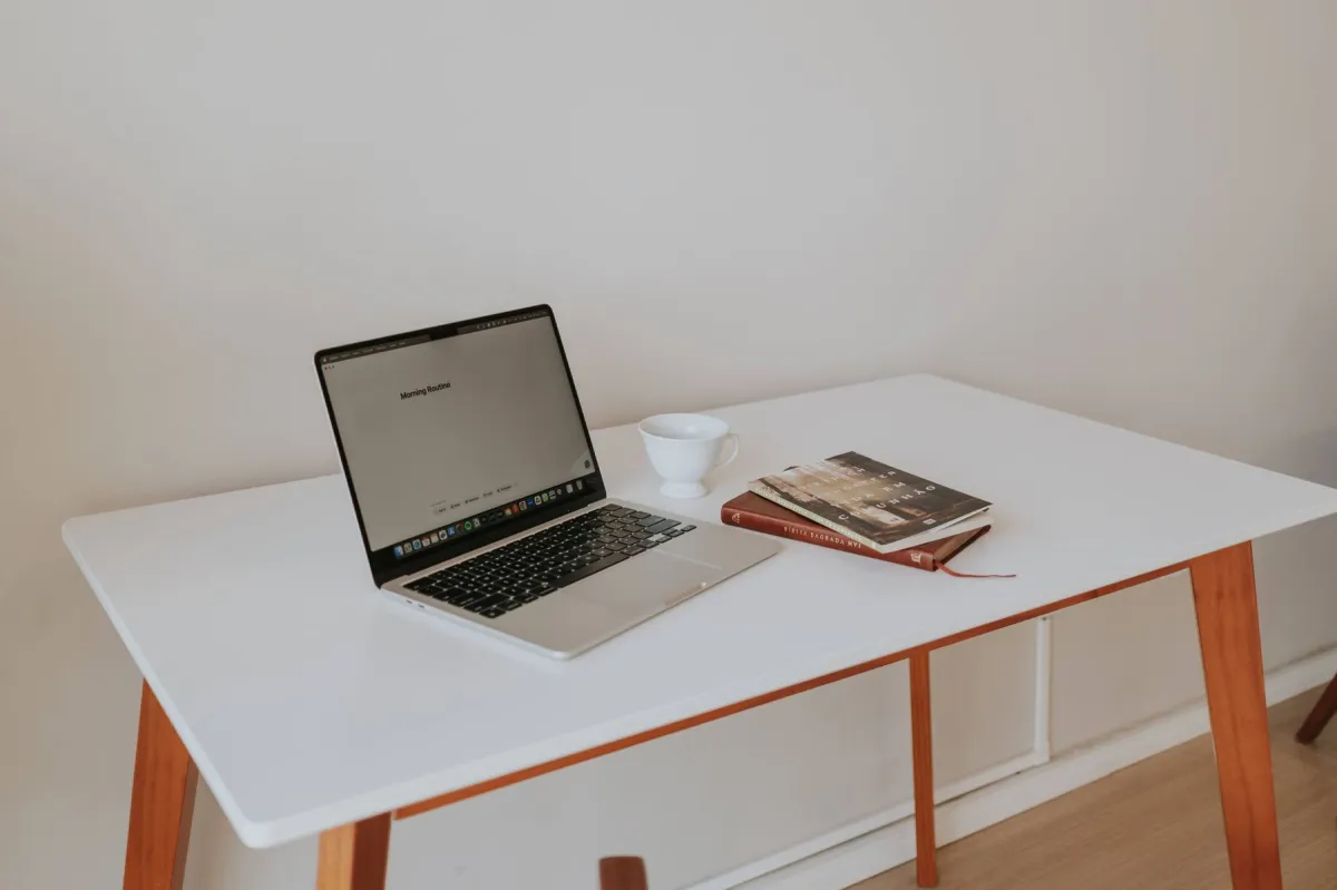Laptop, white cup and two books arranged on a white desk with wooden legs.