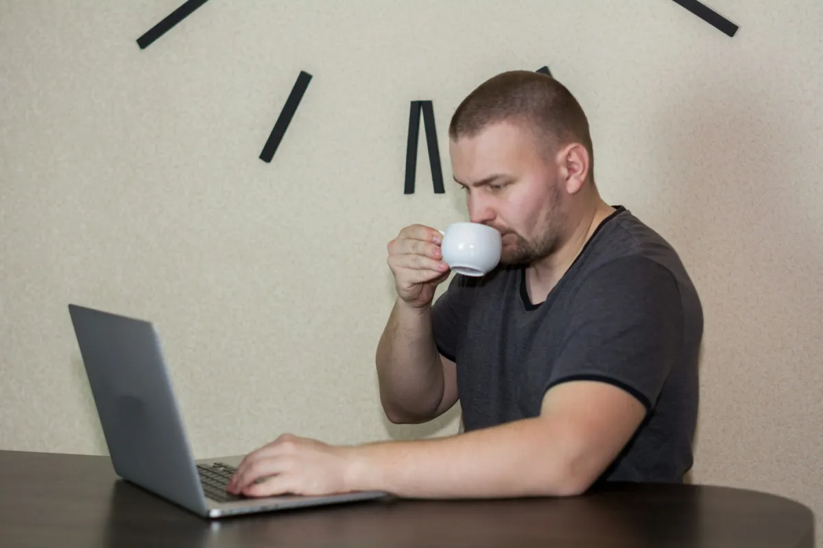 A man drinks from a white cup while using a laptop at a table beneath a large wall clock.