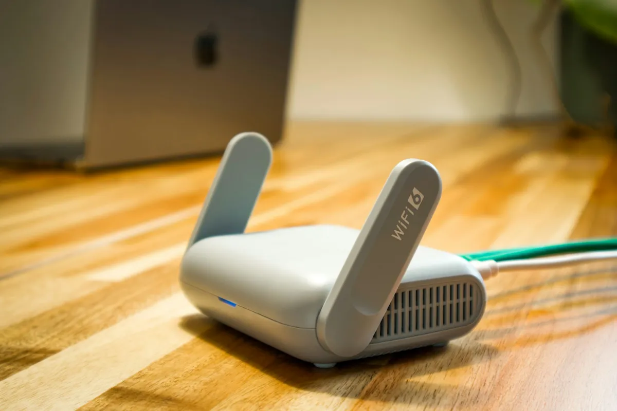 Compact white router with two upright antennas and connected cables on a wooden desk, with a laptop behind it.