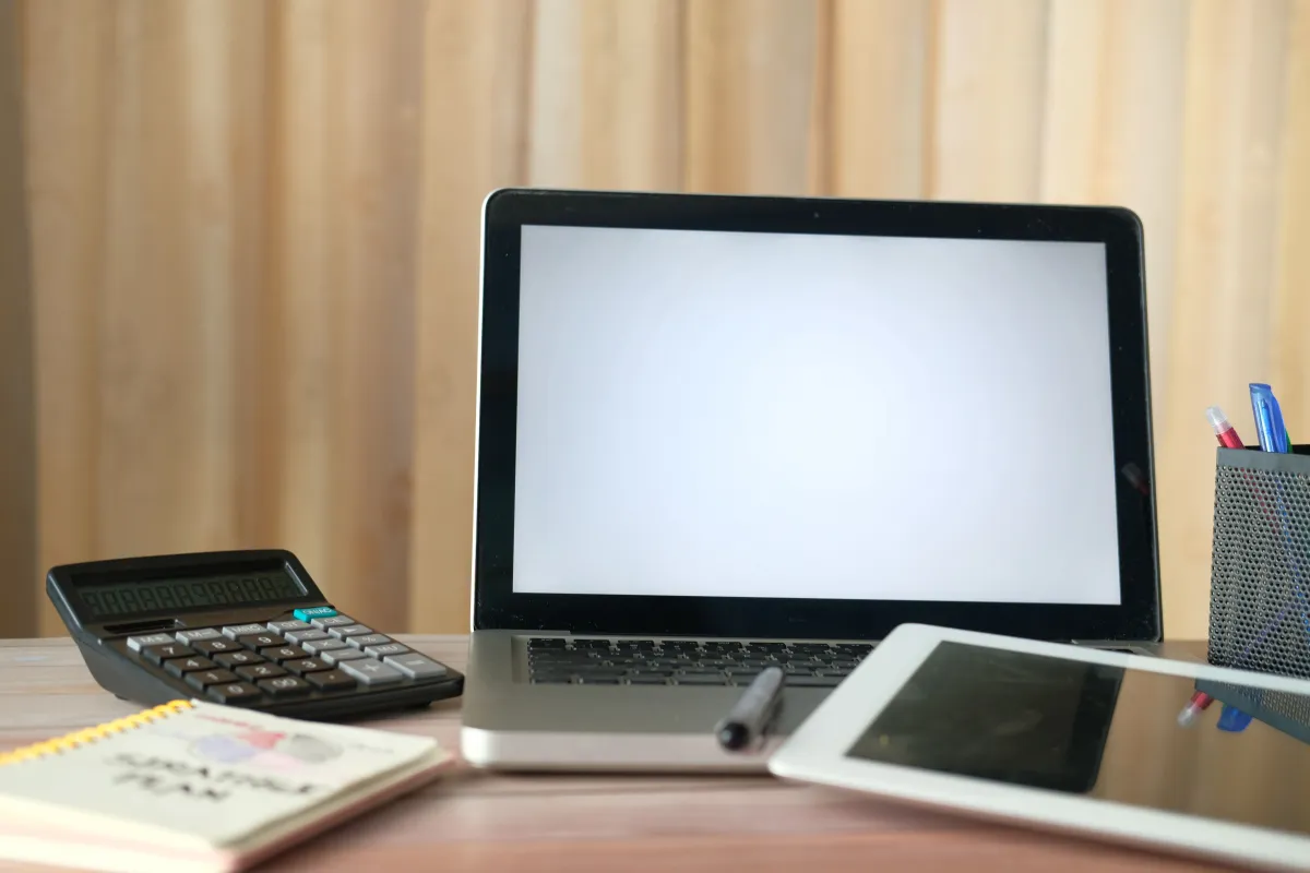 A laptop with a blank screen beside a calculator, tablet and stationery on a wooden desk.