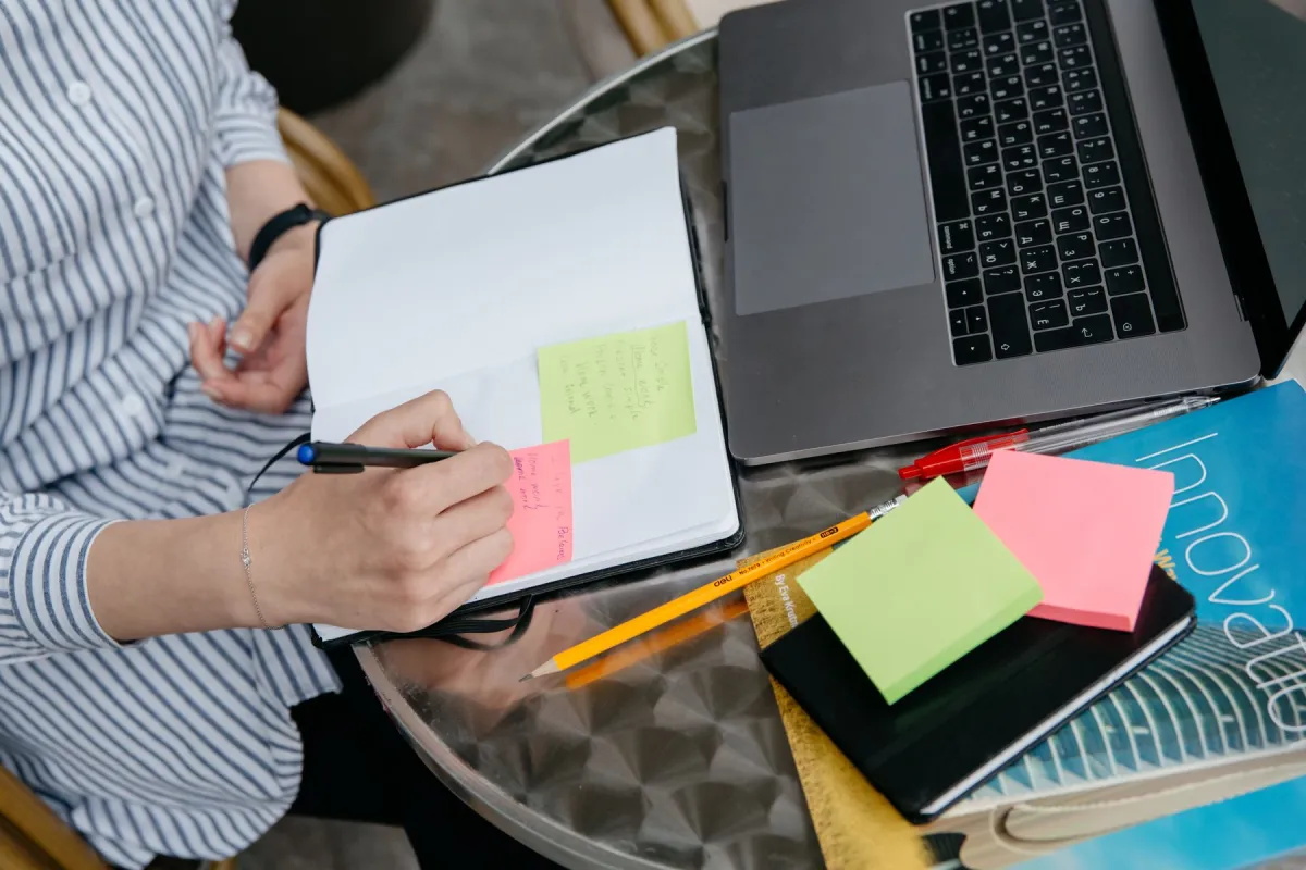 Hands writing notes beside an open laptop and sticky notes