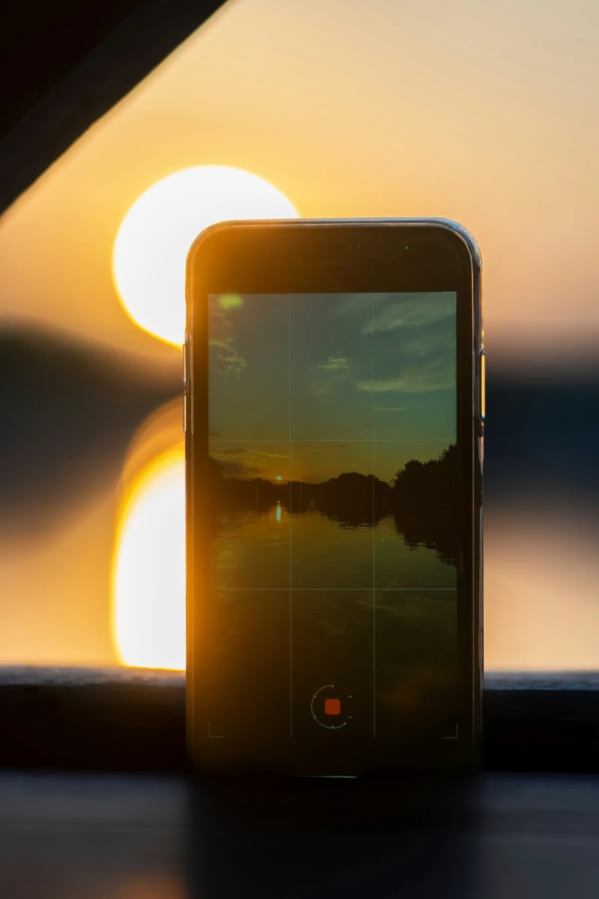 A smartphone standing beside a window shows a sunset in its camera view, with the sun glowing behind it.