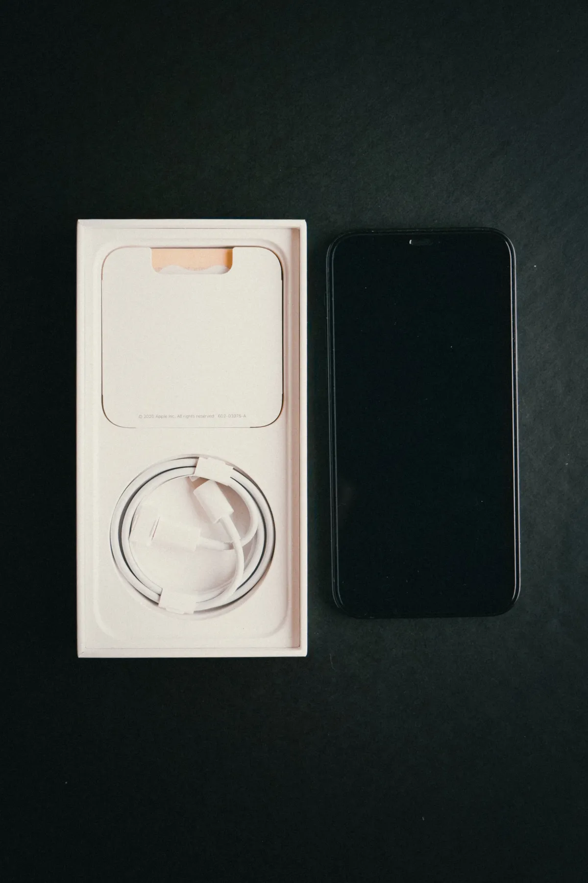 A black smartphone beside an open white box containing a coiled charging cable on a dark surface