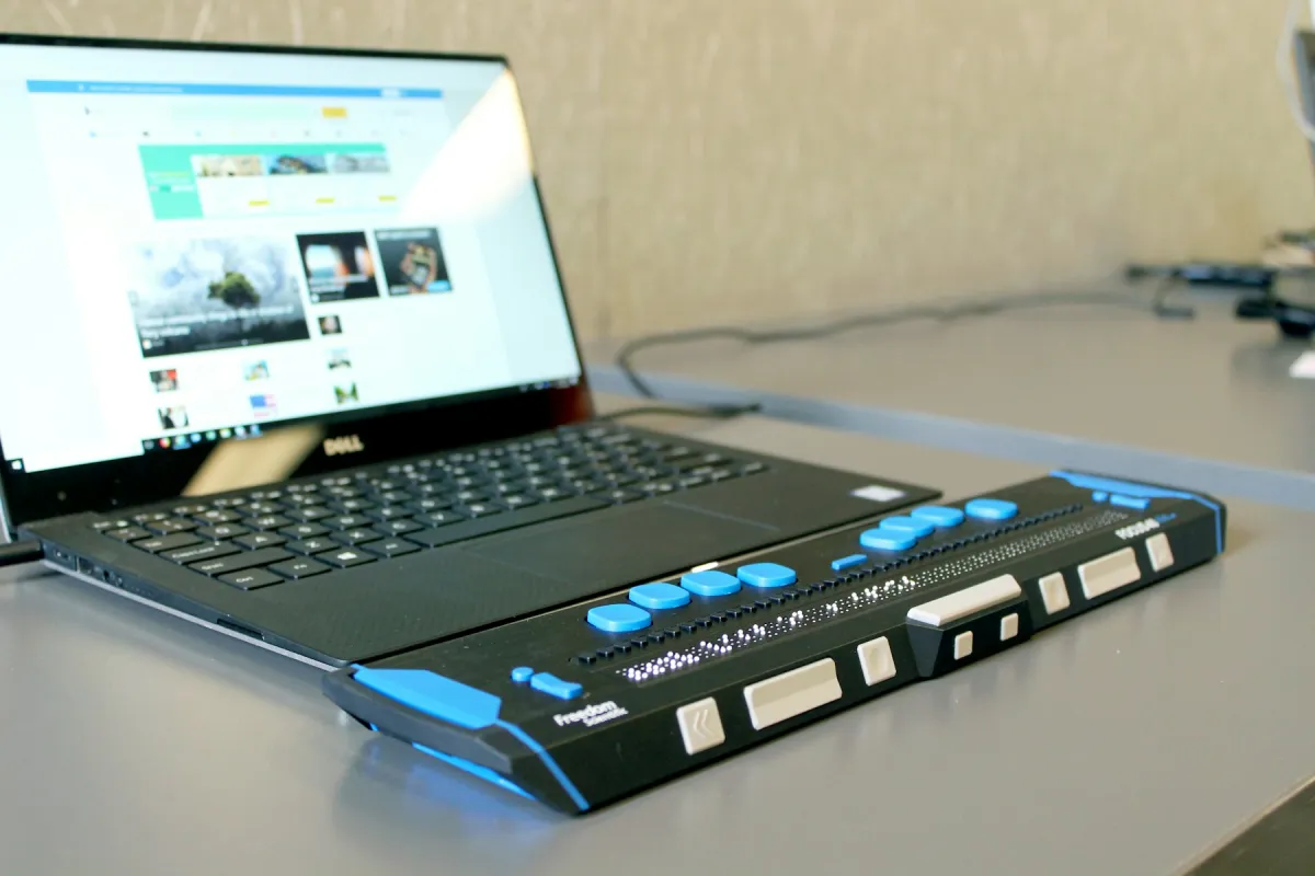 Laptop on a desk with a black refreshable braille display featuring blue buttons in front of its keyboard.
