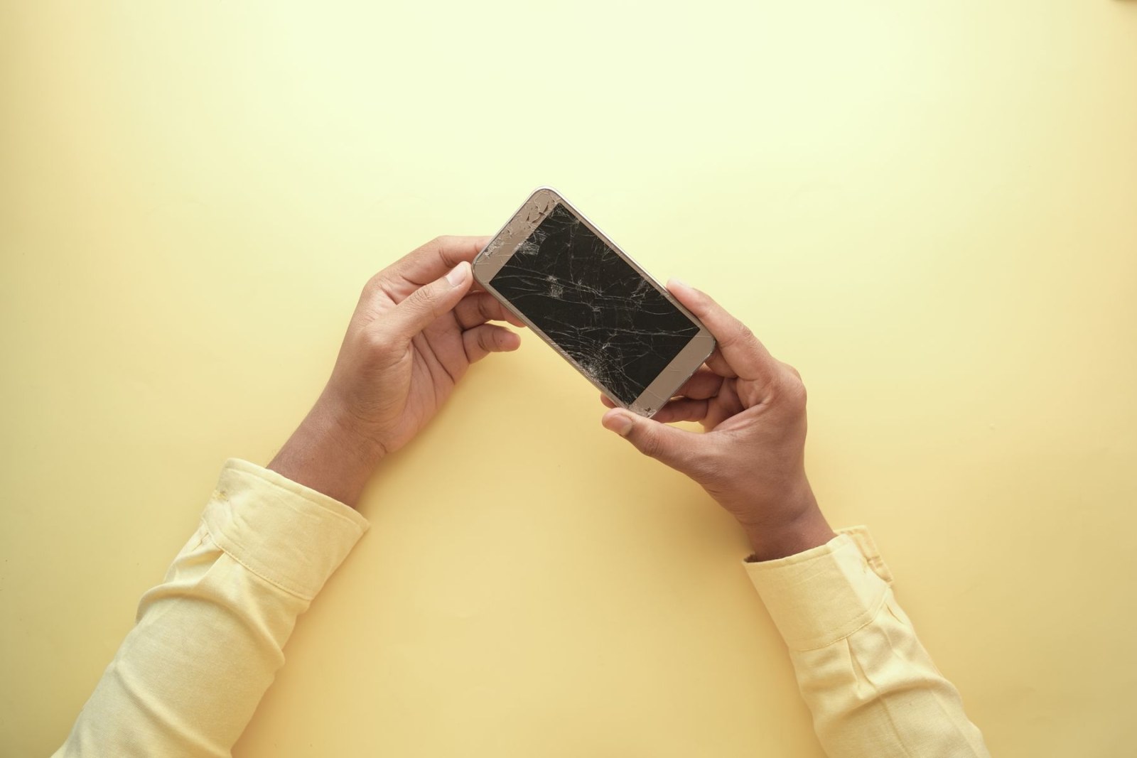 Hands hold a smartphone with dense visible cracks across its front glass against a yellow background.